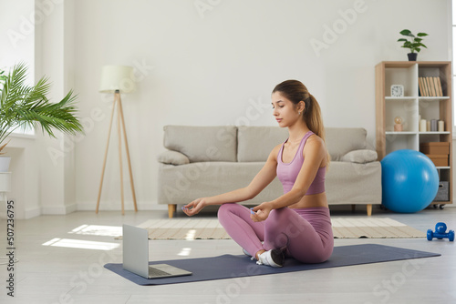 Athletic young woman meditating in front of laptop listening to calm music or affirmations. Woman dressed in comfortable sportswear sits with her eyes closed in lotus position at home in living room.