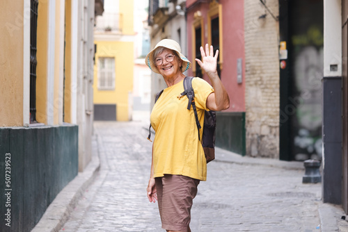 Happy senior woman tourist in yellow t-shirt and cap walking in the alleys of the old town in Seville, Spain looking at camera waving hand
