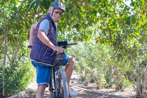 Beautiful active senior man wearing helmet and sunglasses running outdoors in the park with electric bicycle enjoying healthy lifestyle #507239538