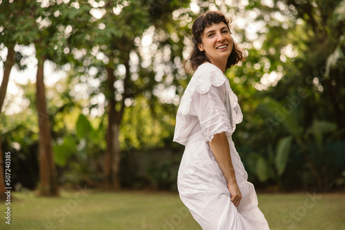 Attractive young caucasian girl is enjoying summer sun on background of trees. Brunette wearing white summer dress is looking at camera. Rest time concept.