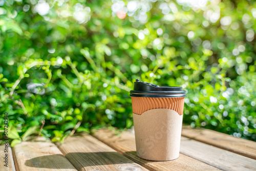 paper coffee cup on the wooden floor, green tree bokeh background in the morning.