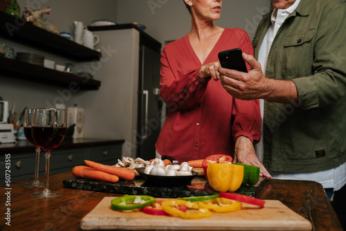 caucasian elderly couple cooking in kitchen looking at cellphone