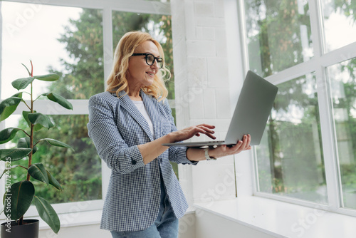 Image of cheerful mature businesswoman standing at office using laptop computer. Portrait of a smiling senior lady holding laptop computer