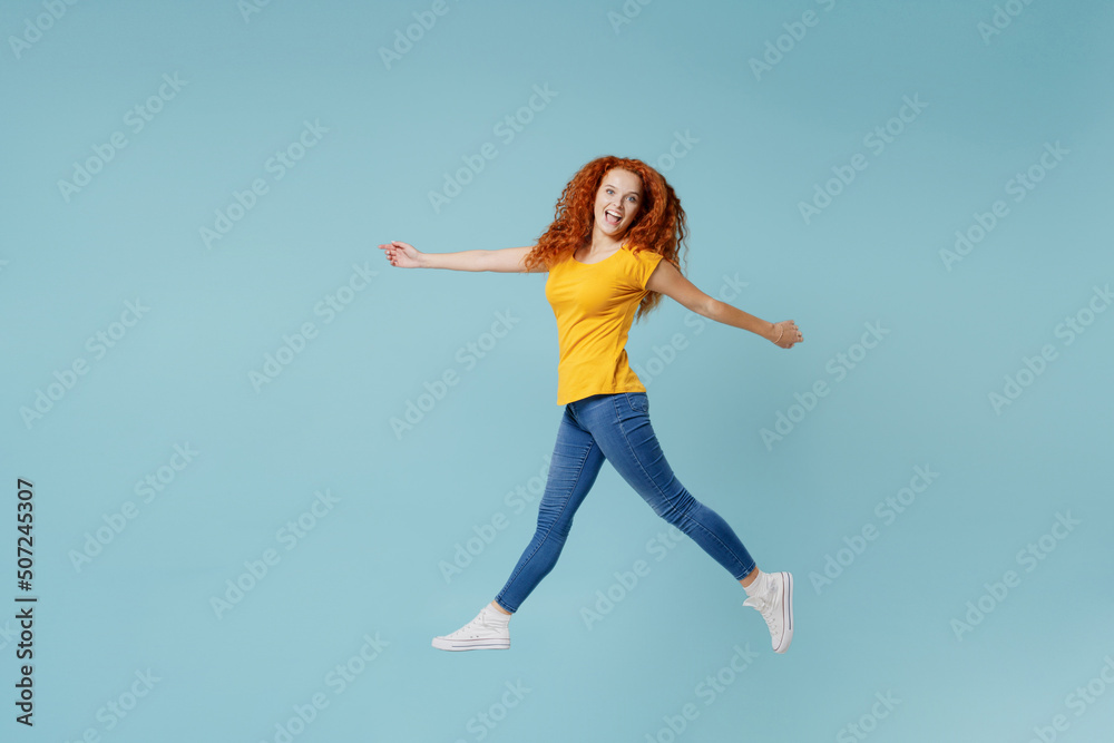 Full body side view young smiling happy redhead woman 20s wearing yellow t-shirt jump high walk go look camera isolated on plain light pastel blue background studio portrait. People lifestyle concept.