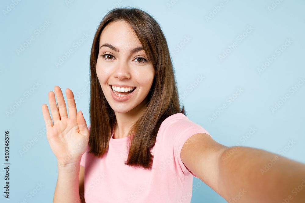 Fototapeta premium Close up young smiling happy woman 20s wear pink t-shirt doing selfie shot pov on mobile cell phone waving hand isolated on pastel plain light blue background studio portrait People lifestyle concept