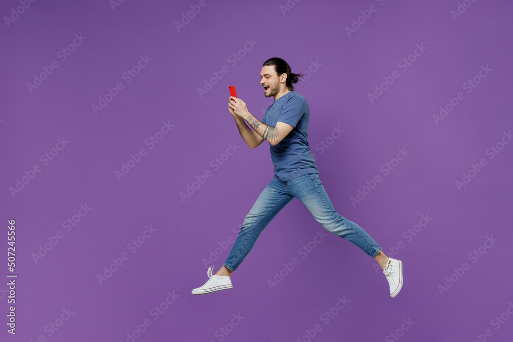 Full body side view young man 20s wearing basic blue t-shirt jump high hold use mobile cell phone hurry up go fast isolated on plain purple color background studio portrait. People lifestyle concept.
