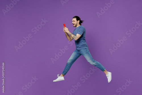 Full body side view young man 20s wearing basic blue t-shirt jump high hold use mobile cell phone hurry up go fast isolated on plain purple color background studio portrait. People lifestyle concept.
