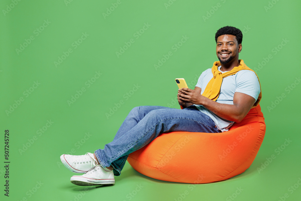 Full size young man of African American ethnicity 20s in blue t-shirt sit in bag chair hold in hand use mobile cell phone isolated on plain green background studio portrait. People lifestyle concept