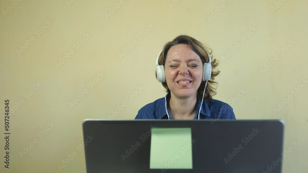 A beautiful forty-year-old blonde woman in white large headphones is talking online on a laptop. Portrait of a fun adult woman in a blue shirt. slow motion A beautiful forty-year-old blonde woman in white large headphones is talking online on a laptop. Portrait of a fun adult woman in a blue shirt. slow motion