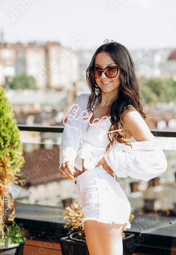 Bachelorette party. Happy future bride in a white shirt and shorts poses on the roof of the house overlooking the city.