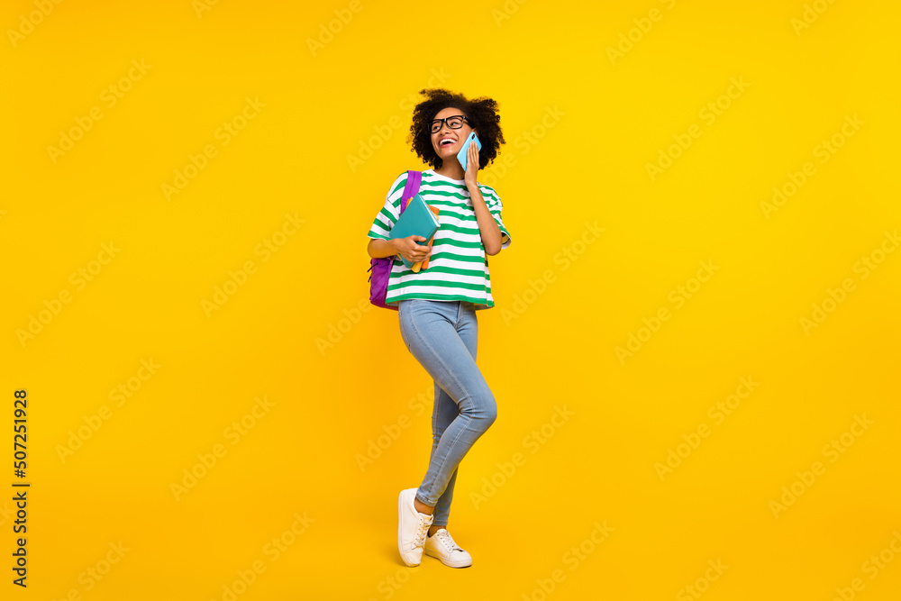 Full size photo of impressed millennial lady tell telephone hold book look promo wear glasses t-shirt bag jeans shoes isolated on yellow background