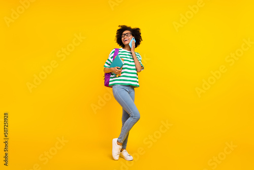 Full size photo of impressed millennial lady tell telephone hold book look promo wear glasses t-shirt bag jeans shoes isolated on yellow background © deagreez