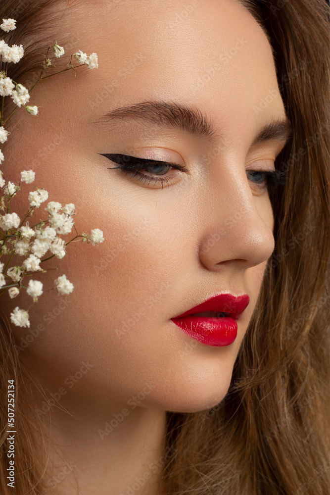 Fototapeta premium Close up portrait of a face with beautiful skin and evening makeup, eyeliner and long eyelashes, puffy red lips and white flowers. Fashion photography. Cosmetology and spa. Freshness of spring