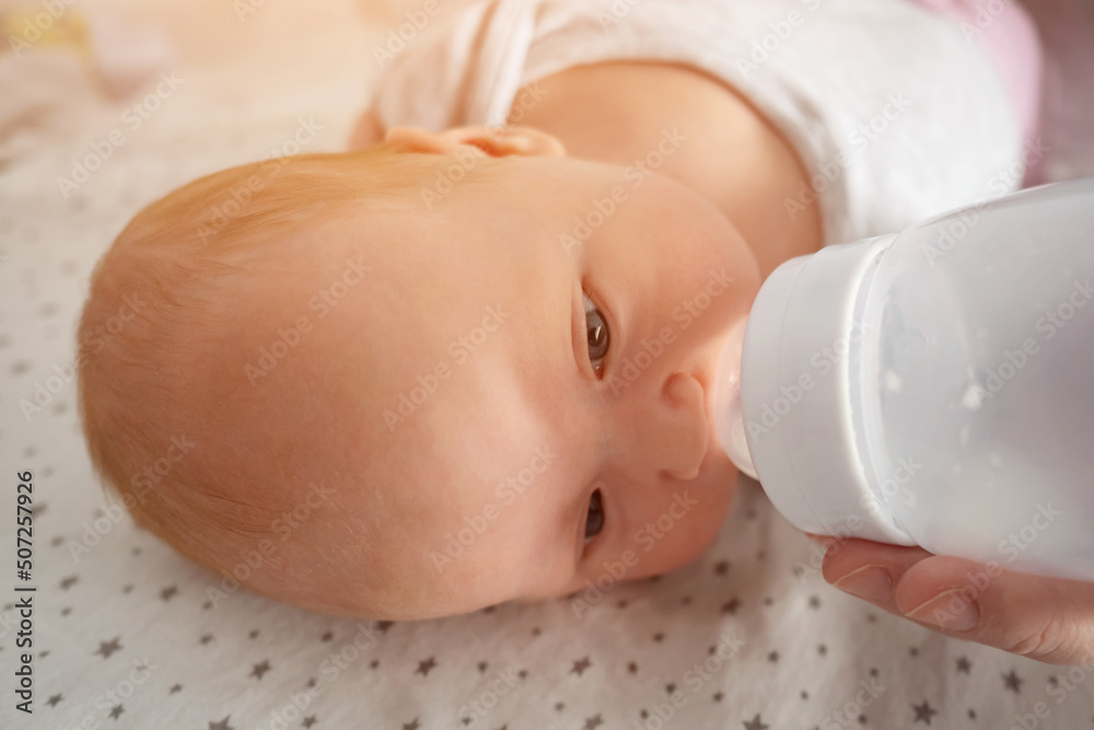 Newborn baby girl with half closed eyes drinks water from plastic bottle lying on bed. Mother feeds cute little kid at home closeup, side view Newborn baby girl with half closed eyes drinks water from plastic bottle lying on bed. Mother feeds cute little kid at home closeup, side view
