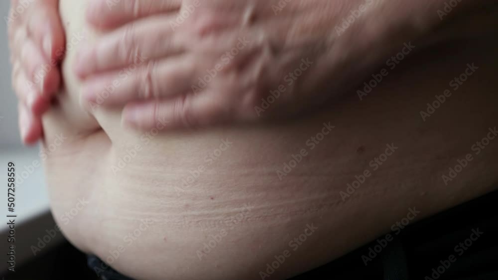 Woman strokes and squeezes fat belly. Aged lady measures fat on stomach near windowsill. Plus-size woman hands hold fat belly with flabby sagging skin Woman strokes and squeezes fat belly. Aged lady measures fat on stomach near windowsill. Plus-size woman hands hold fat belly with flabby sagging skin