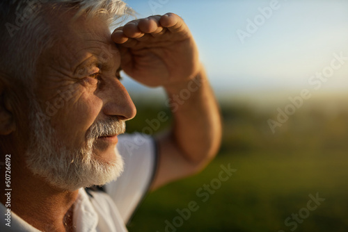 Senior bearded man using hand to protect eyes from sun, smiling outside. Close up view of male tourist squinting from bright light, while looking into distance, during mountain trip. Concept of watch. photo