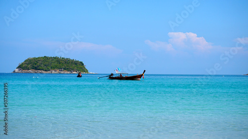 A lonely island in the sea. A Thai fishing boat floats nearby. Clear blue water, snow-white sand. Small clouds in the sea. Palm trees and trees grow on the island. Beautiful Kata beach, Phuket © SergeyPanikhin