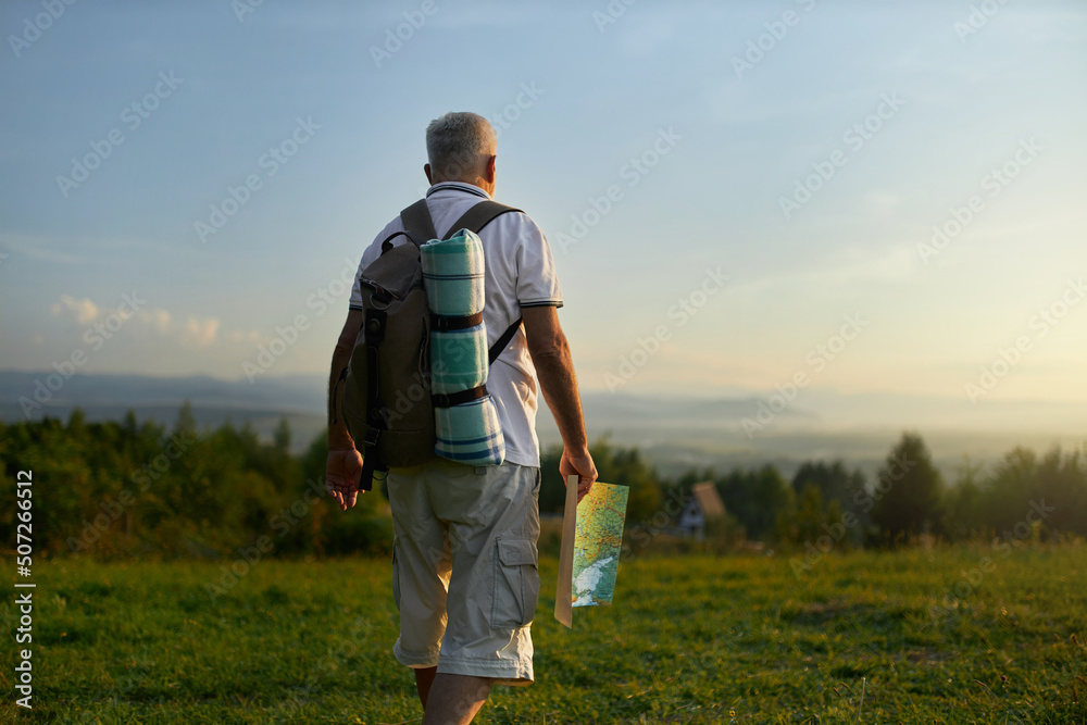 Middle aged man walking on green meadow, while hiking in mountains. Back view of adult male traveler holding map, while backpacking on rural area in summer, with copy space. Concept of backpacking.