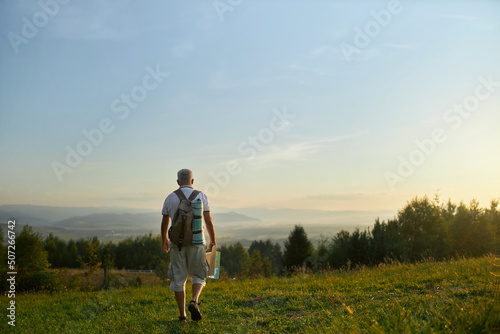 Adult male traveler backpacking in mountains at sunset, with landscape on background. Back view of middle aged man walking down hill, while hiking in summer, with copy space. Concept of backpacking.