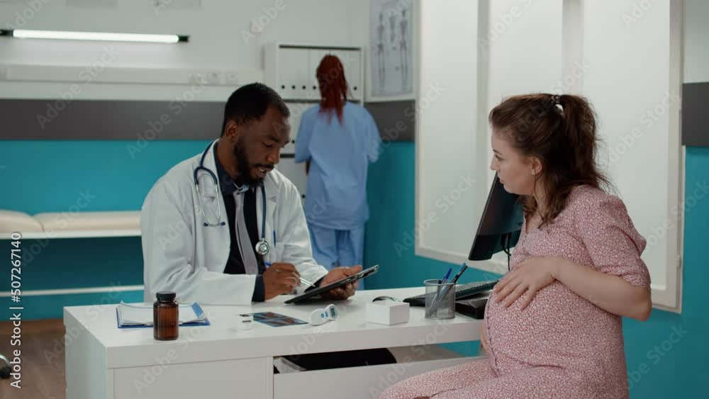 Maternity specialist taking notes on digital tablet at checkup with expectant woman to give prescription medicine. Male physician talking to patient with pregnancy belly at appointment. Maternity specialist taking notes on digital tablet at checkup with expectant woman to give prescription medicine. Male physician talking to patient with pregnancy belly at appointment.