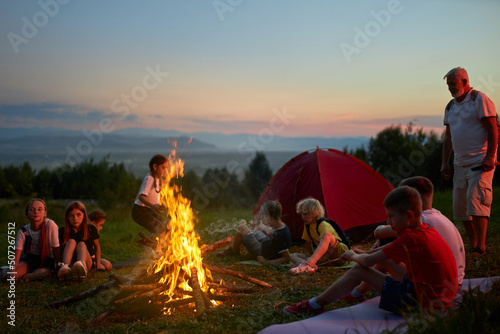 Happy children with adult instructor sitting around campfire in evening. Side view of group of teen tourists resting, having fun, with scenic mountain landscape on background. Concept of travelling.