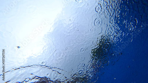 View from under the water to the sky and rain. Drops fall on the surface of the water create water circles. Gray clouds and blue sky are visible. Dark green highlights of trees. An unusual look