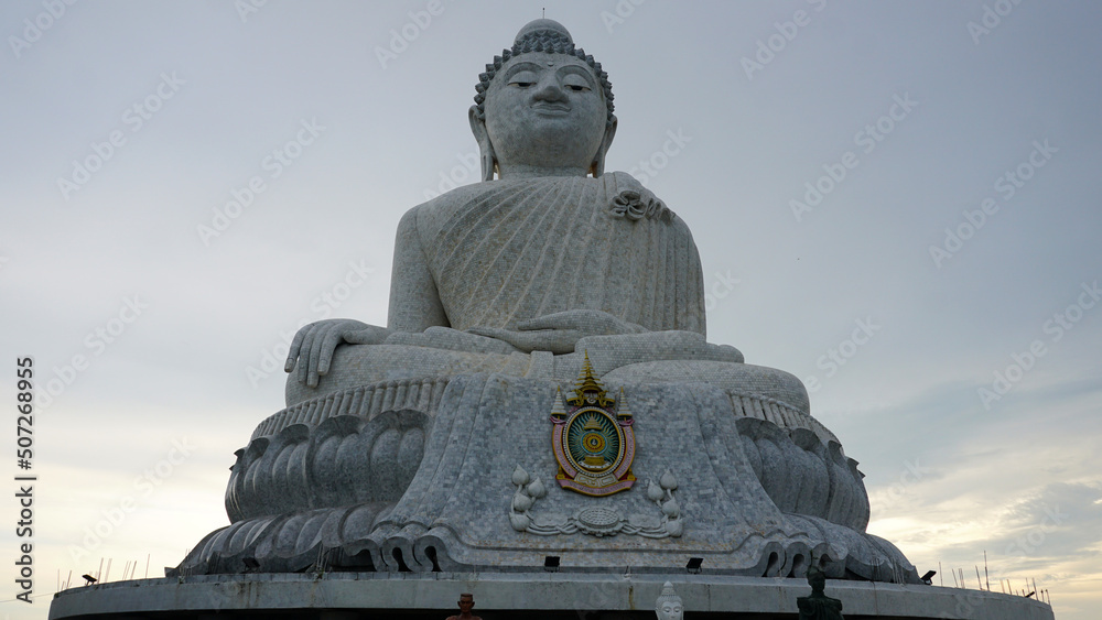 Fototapeta premium The Big Buddha looks straight into the eyes (into the camera). He sits in the lotus position and looks down at people. There is a temple in the center. Gray clouds. Buddha statue in Phuket, Thailand