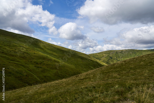 Rolling grassy hills under cloudy blue sky in autumn season. Panoramic view of green mountain ridge outdoors in overcast day, with copy space. Concept of landscape. 