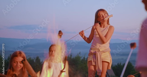 Front view of pretty blond teenage girl eating marshmallow, roasted on fire in mountains. Female kid standing near fire, enjoying roasted meal, with group of kids at campsite. Concept of travelling.