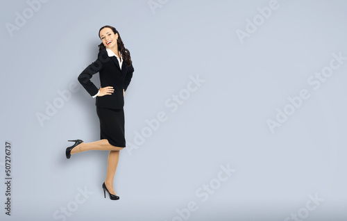 Full body length of happy excited smiling businesswoman with hands on hips, copy space for text. Confident woman in black suit standing dancing on grey background. Business concept.