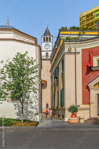 Tirana, Albania - June 21, 2021: View of the Clock Tower between the buildings of Ethem Bey Mosque and Tirana City Hall. Ancient architecture in the center of the Albanian capital