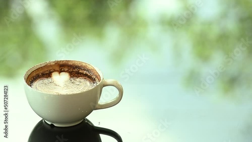 Latte art coffee cup on glass table. Blurred leaf reflection background.