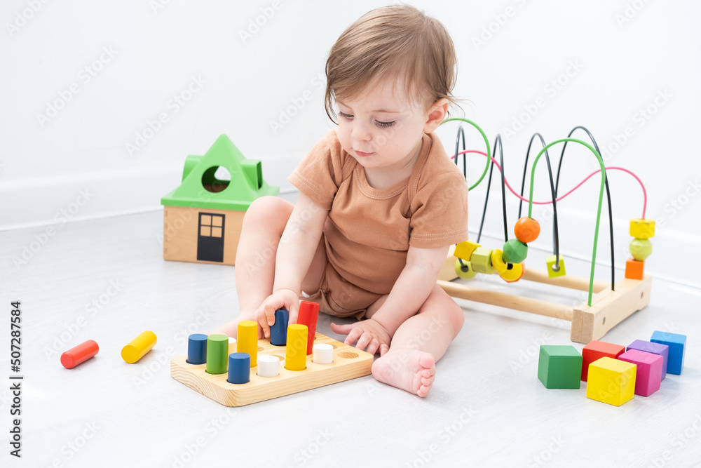 cute baby girl in brown bodysuit playing with colorful wooden toys on white room cute baby girl in brown bodysuit playing with colorful wooden toys on white room