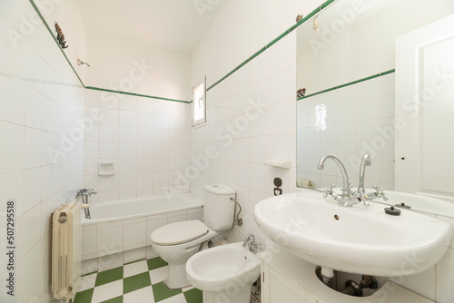 Bathroom with white wooden furniture with porcelain sink  frameless mirror on the wall  long tub in the background  green upper border and white and green ceramic floors