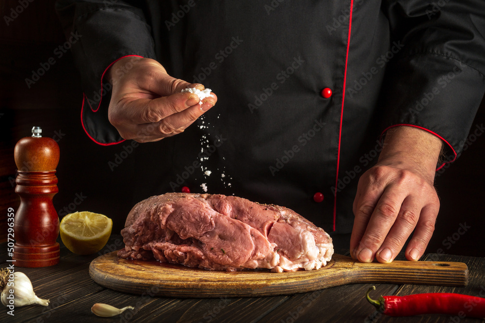 Chef sprinkles raw meat with salt. Preparing fresh meat before roasting. Menu concept for restaurant or hotel