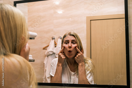 Second chin, obesity. Portrait of 30 year old girl lifting her cheeks up using her fingers while looking at mirror in bathroom. Aging and appearance problems.. Facelift, plastic surgery, facebuilding photo