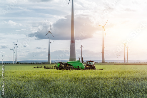 Scenic landscape view big modern tractor machine with sprayer equipment spraying fertilizer in rapeseed agricultural field against windfarm power windmill. Arable land cultivation sustainable energy