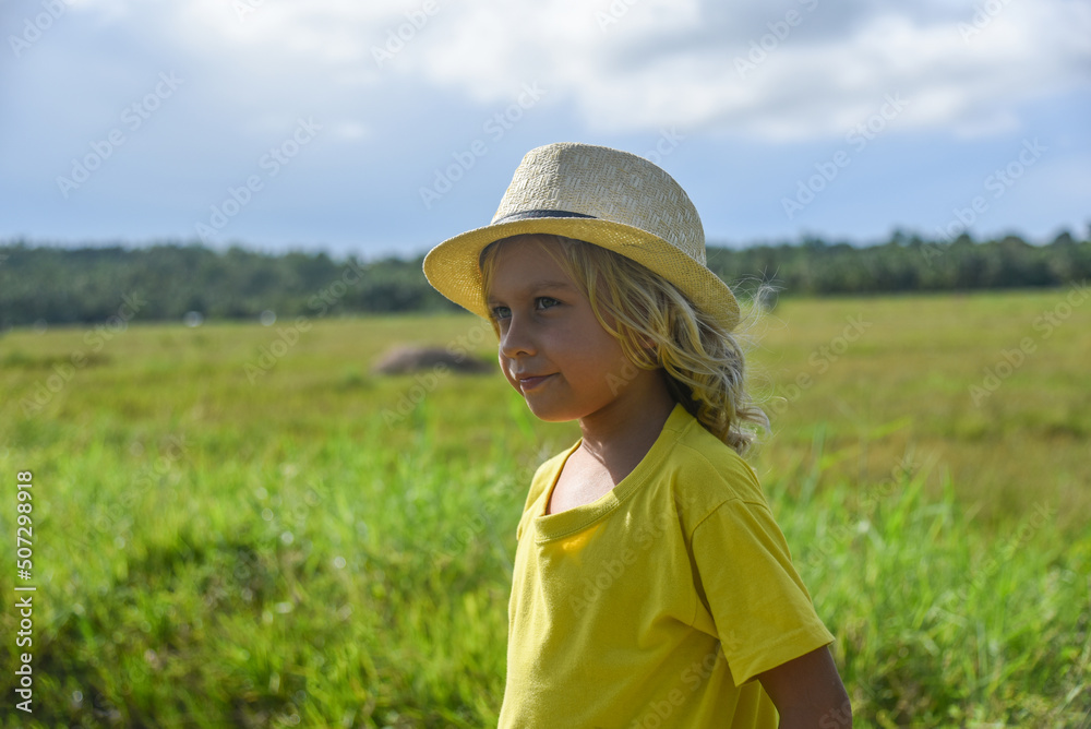 A little curly-haired boy walks in nature in a hat and a yellow T-shirt. Family vacation, traveler, trip, summer time. Green grass and blue sky in the background, front view, life style