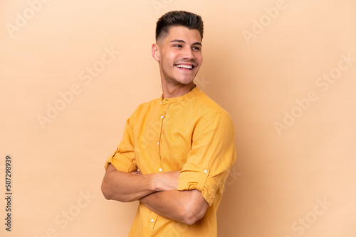 Young caucasian man isolated on beige background with arms crossed and happy