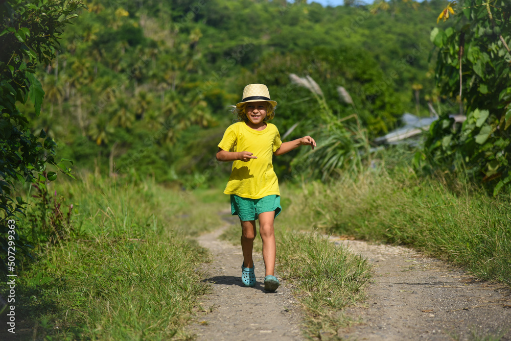 A little curly-haired boy runs in nature in a hat and a yellow T-shirt. Family vacation, runner, trip, summer time. Green grass and blue sky in the background, front view, life style