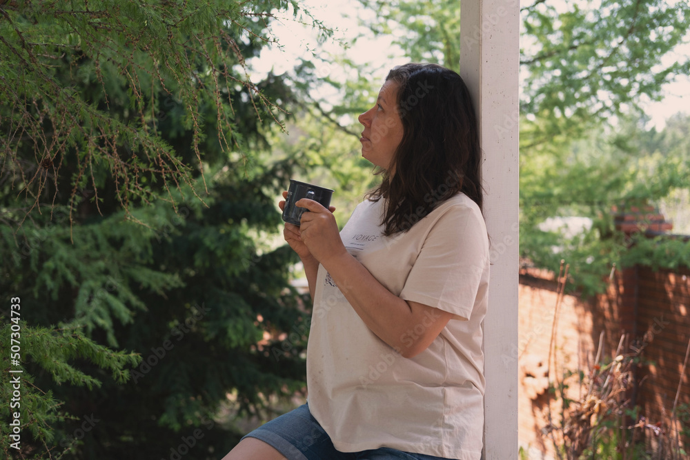 A romantic portrait of a beautiful young woman in a T-shirt and jeans, resting on a balcony overlooking the forest, with a cup of tea or coffee in her hands. A romantic portrait of a beautiful young woman in a T-shirt and jeans, resting on a balcony overlooking the forest, with a cup of tea or coffee in her hands.