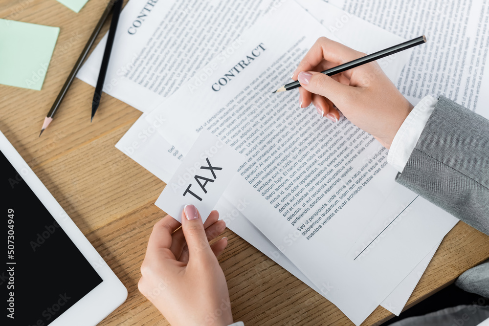 cropped view of tax inspector holding paper with tax lettering while checking contract near digital tablet. cropped view of tax inspector holding paper with tax lettering while checking contract near digital tablet.
