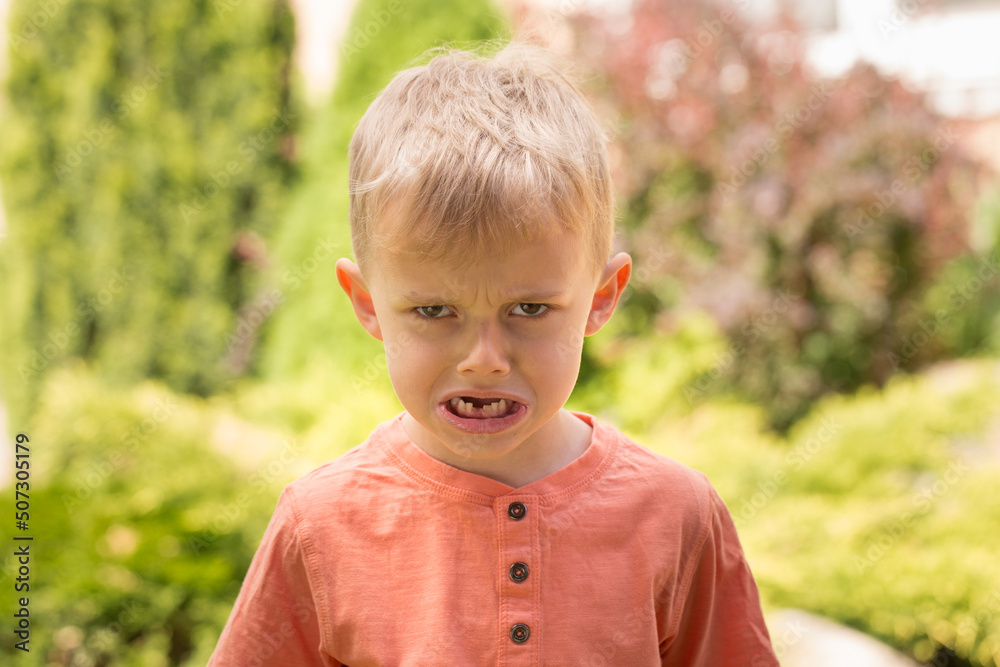 five-year-old boy making faces. A five-year-old blond boy makes faces while standing on the street in sunny weather five-year-old boy making faces. A five-year-old blond boy makes faces while standing on the street in sunny weather
