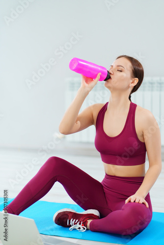 a young woman drinks water in a burgundy tracksuit with laptop on a mat in a gym