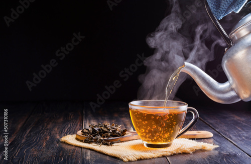 Pour a hot herbal teapot into a glass cup, teacup, and dry tea leaves in a wooden spoon and place it on a black wooden table against a dark background.