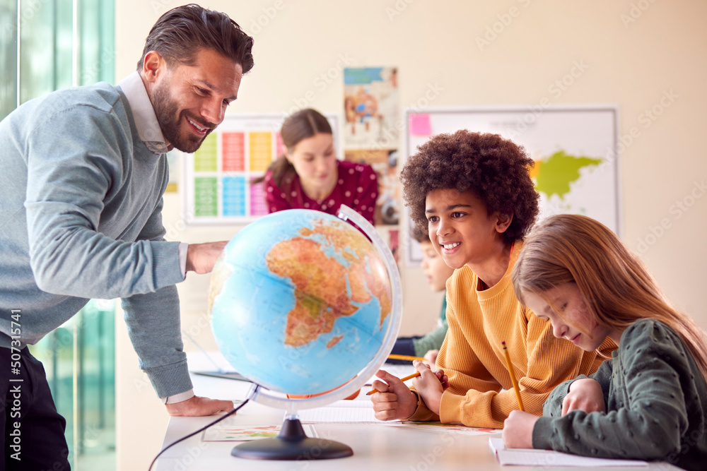 Group Of Multi-Cultural Students With Teachers In Classroom Looking At Globe In Geography Lesson Group Of Multi-Cultural Students With Teachers In Classroom Looking At Globe In Geography Lesson