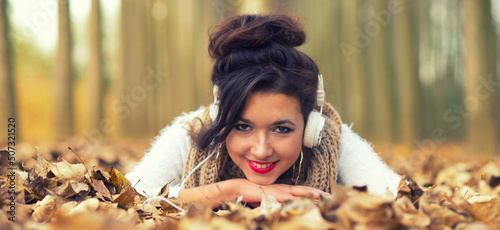 Young smiling woman lying in autumn leaves and listening music through earphones 