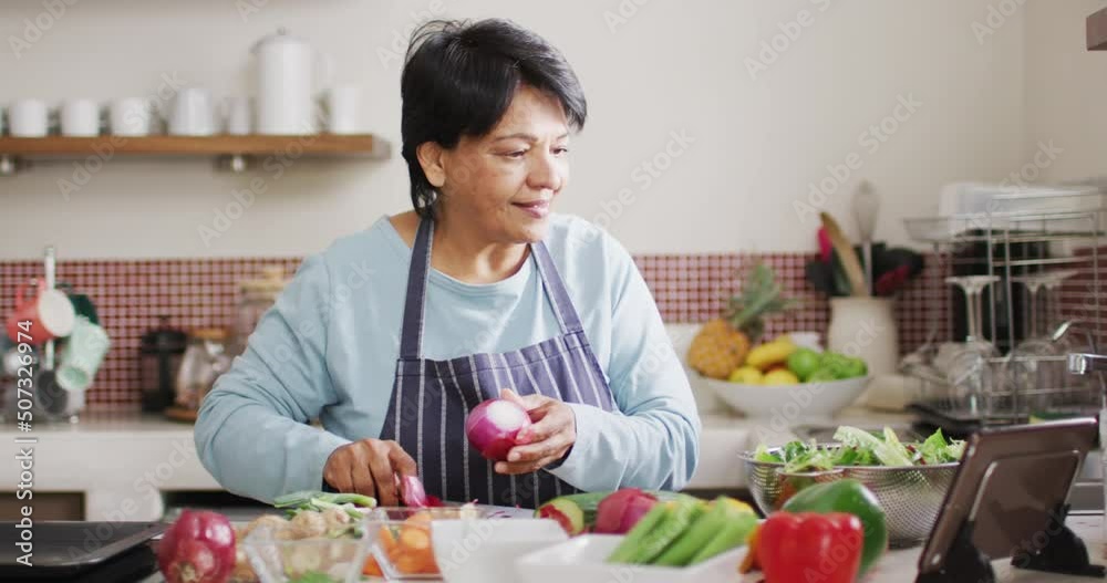 Asian senior woman using digital tablet while removing onions skin in the kitchen at home