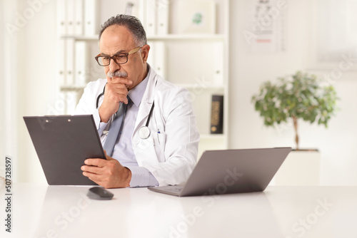 Mature male doctor sitting in an office and reading a document at a desk with a computer