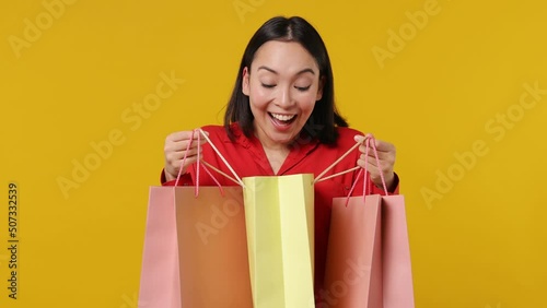Satisfied smiling vivid young woman of Asian ethnicity 20s years old wears red shirt holding looking into package bags with purchases after shopping isolated on plain yellow background studio portrait photo